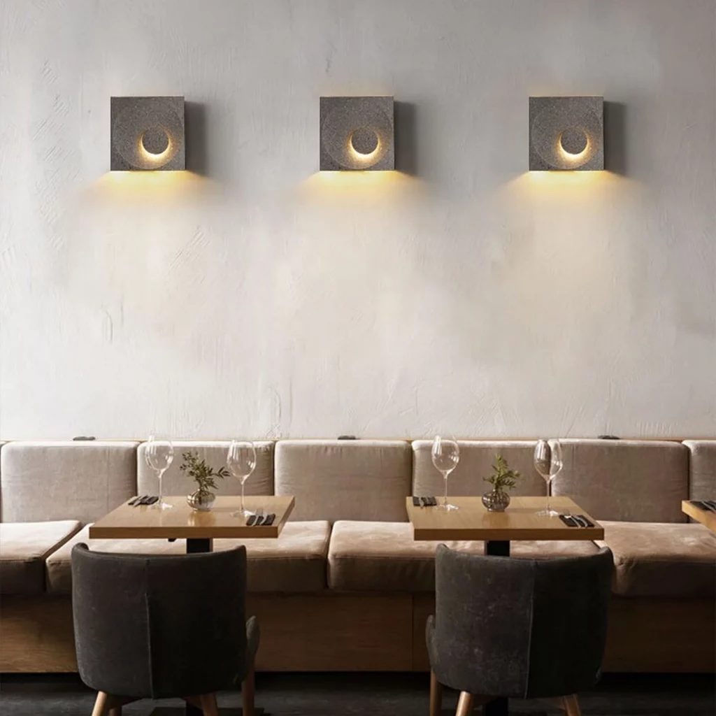 Postmodern Cement Wall Lamp casting a warm crescent glow on a textured wall above a cozy dining seating area with tables and chairs.