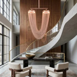 Modern hotel lobby or atrium with a sweeping curved staircase, glass railing, and a large pink multi‑strand chandelier above a black marble table and two cream chairs.